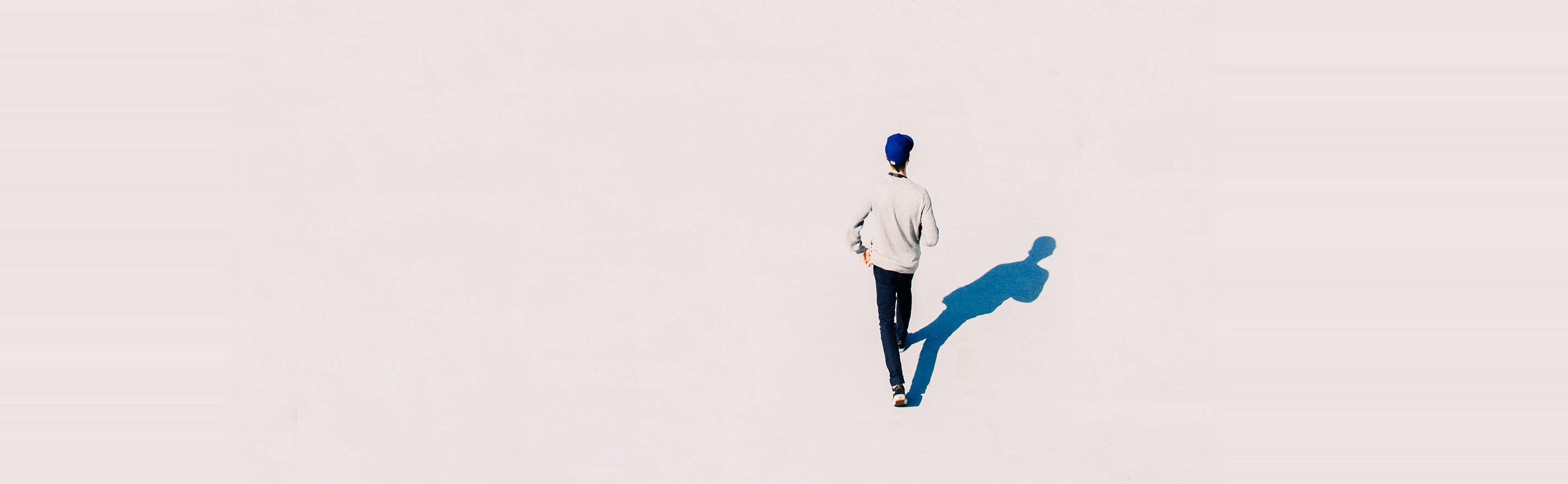 A lone figure walking across a wide pale expanse, casting a long blue shadow